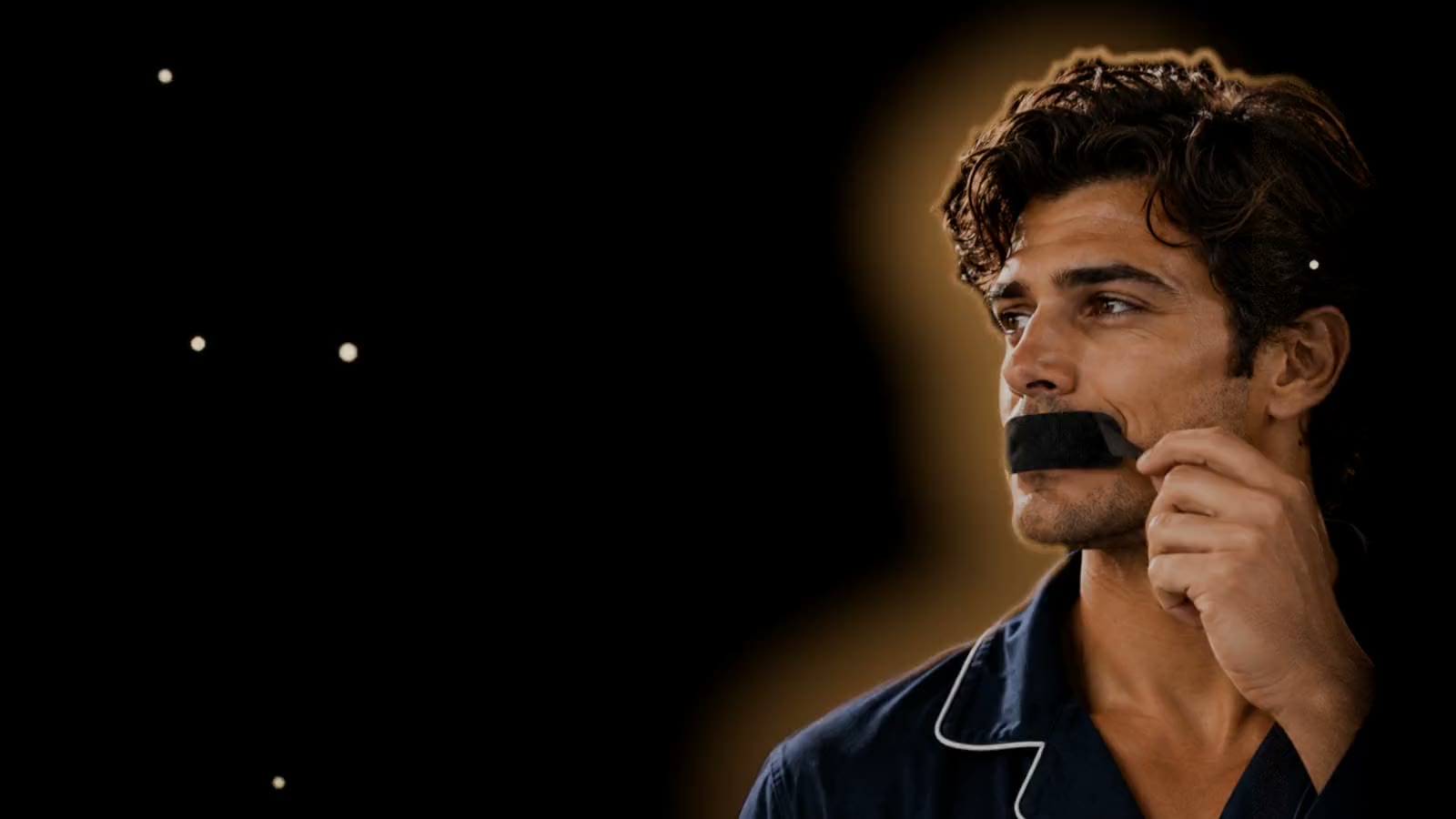 Homem de pijama azul-marinho segurando a Somnus Tape, fita bucal preta, junto aos lábios